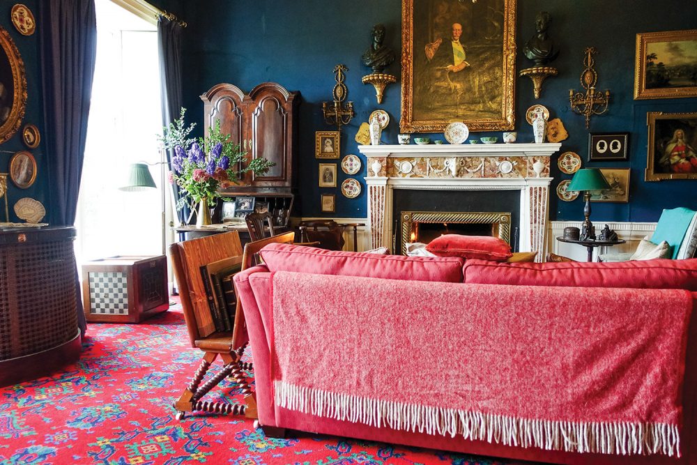 The library features dark blue walls and draperies; a plush, cherry red sofa with a matching fringed throw draped over the back, a bright red fine rug with a geometric pattern of bright teal, blue, and yellow; an ornate fireplace beneath a historical oil portrait in an elaborate gilt frame; and antique furniture and decor.
