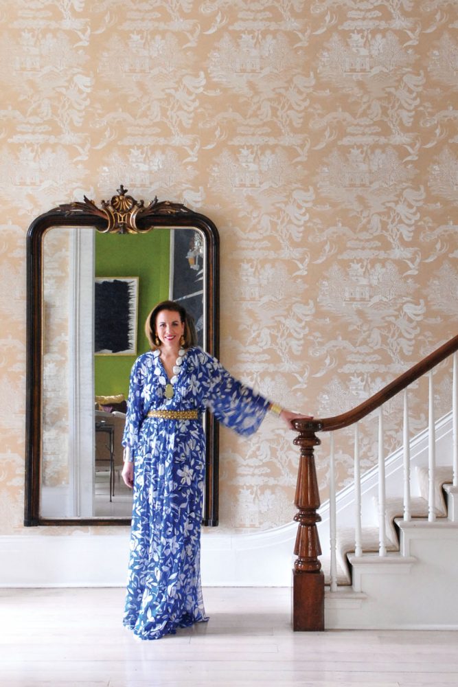 founder of Leontine Linens in a long, blue Oscar de la Renta dress with white flowers, stands at the bottom of an elegant staircase 