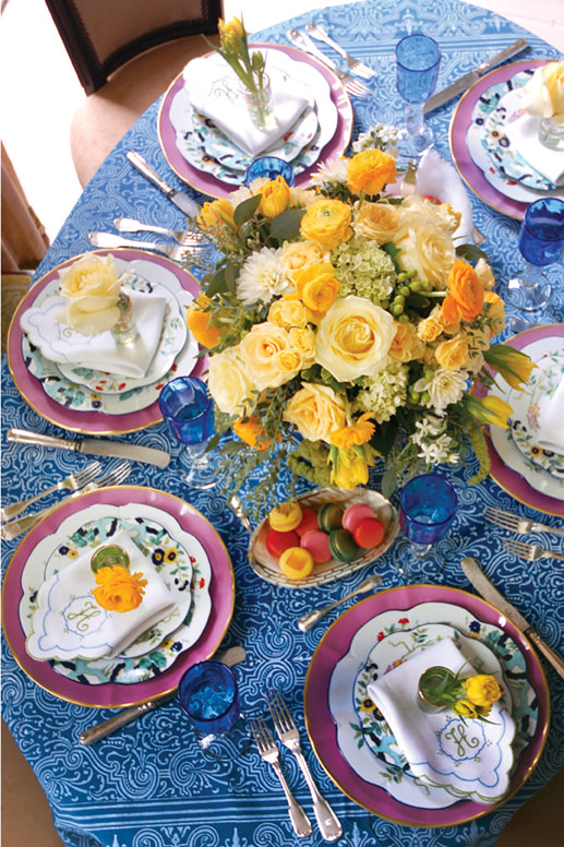 A cheerful table setting features a blue batik-print tablecloth, yellow flowers, pink chargers by Anna Weatherley, ‘Paradis Bleu’ plates by Royal Limoges, and scalloped white napkins with green and blue embroidery