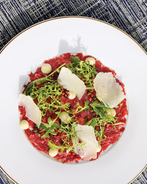 Carpaccio artfully plated in a white-brimmed white plate, garnished with fresh herbs and shaved cheese
