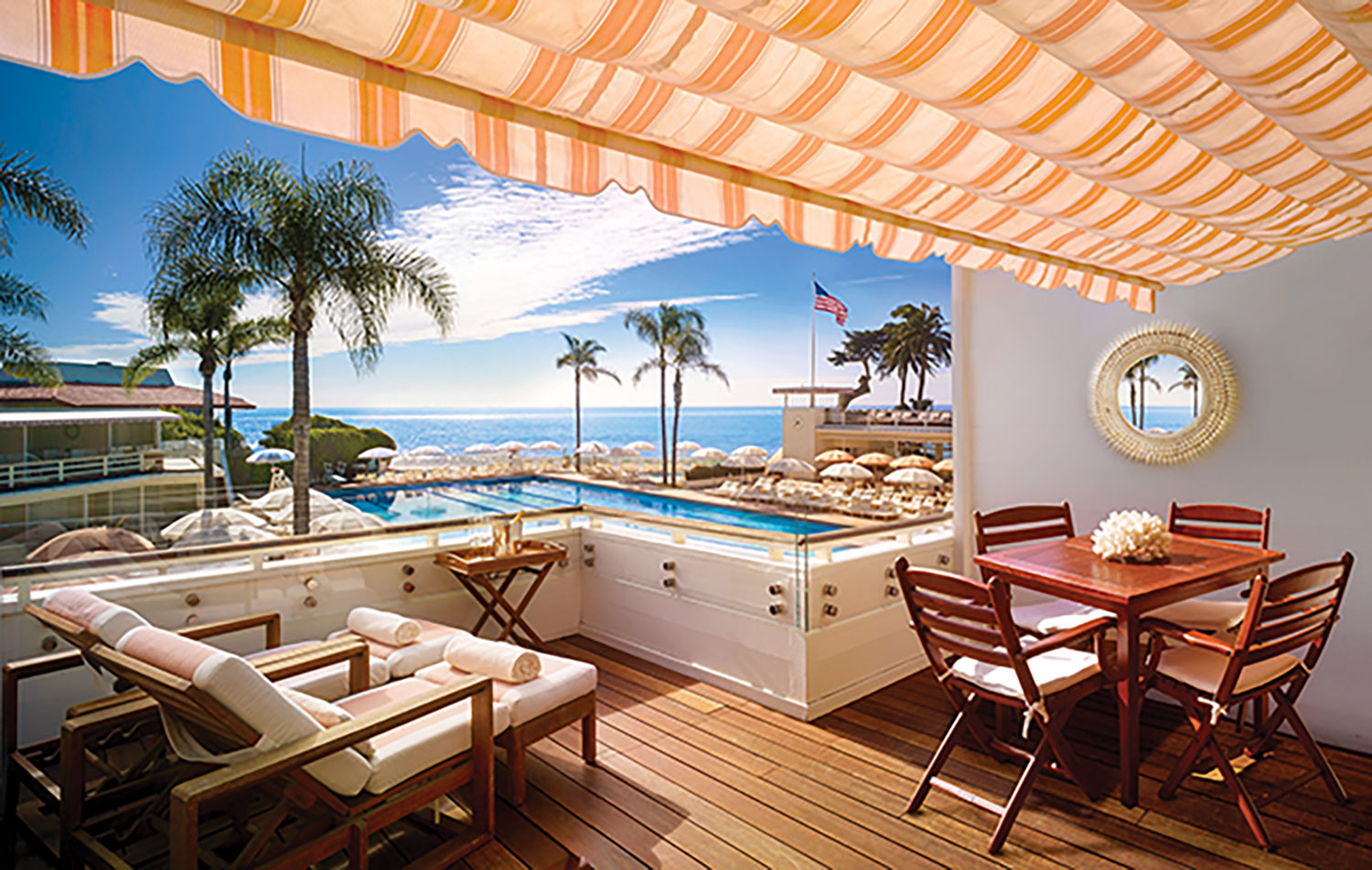 A covered patio area, featuring wood plant floors, teak patio furniture, and a yellow and white striped awning, looks out onto a pool at the Four Seasons Hotel Santa Barbara and a view of the coast beyond
