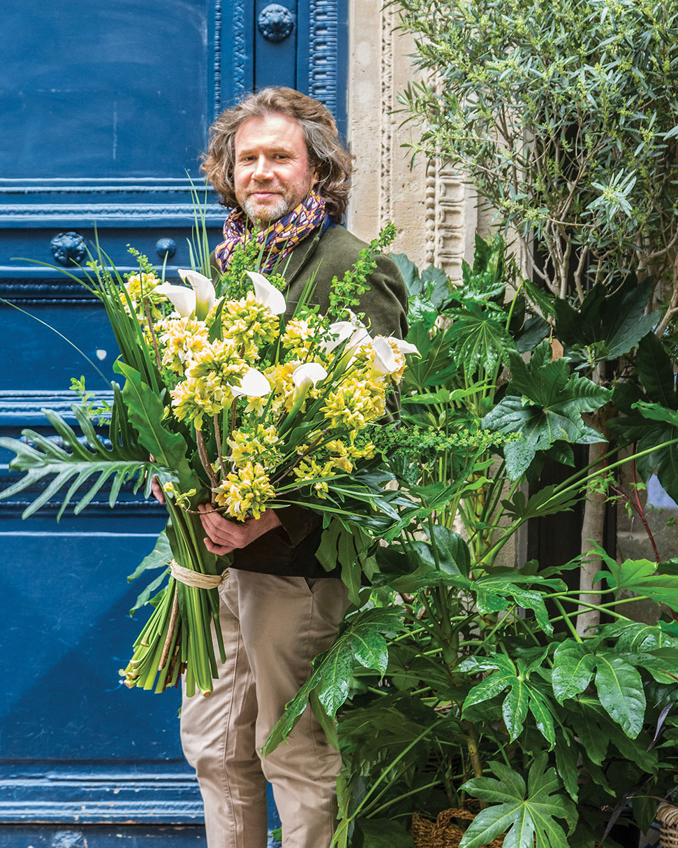 Parisian florist Stephane Chapelle stands against the bright blue painted door of this French flower shop, holding a massive bouquet of tropical yellow flowers and foliage.