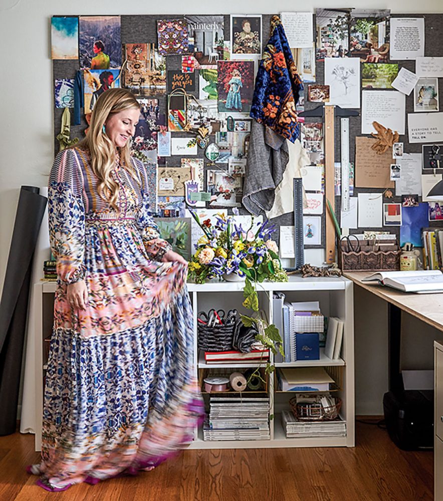 Artist Marcy Cook wearing a long Boho-style dress stands in front of her inspiration board in her studio