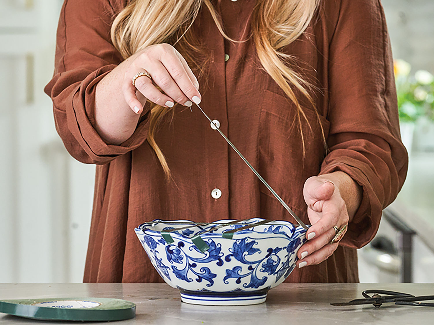 March Cook, dressed in a deep brown button-up, creates a grid of floral tape over a blue-and-white bowl