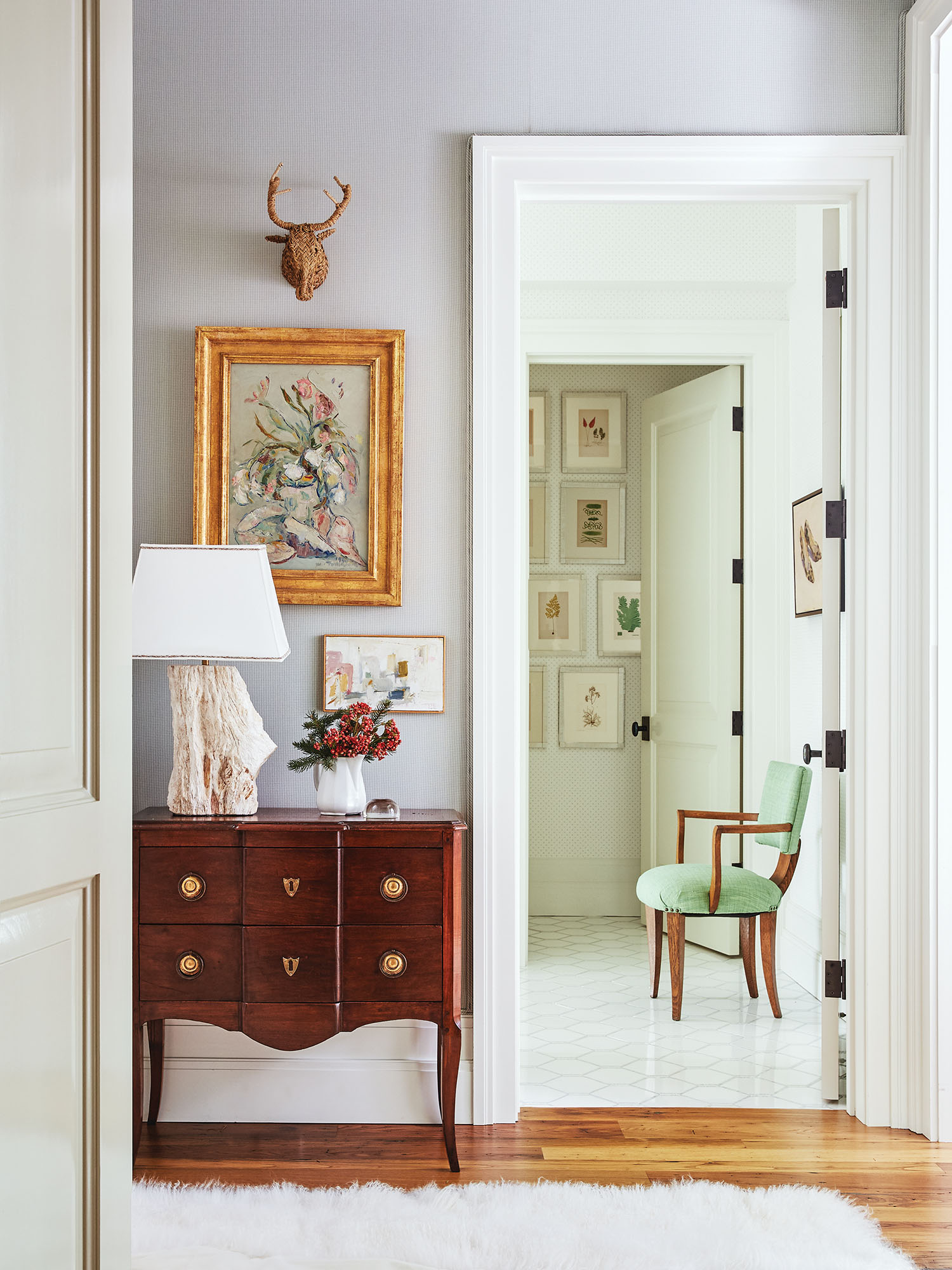 A bright hallway has a fur rug, an antique side table, a floral painting, and a green chair.