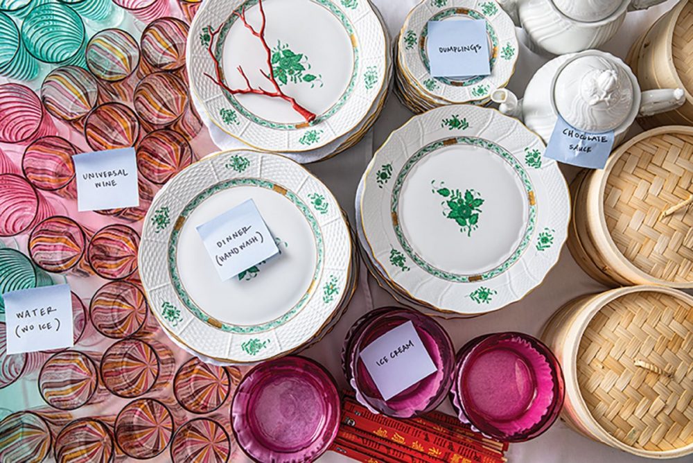 A table full of colorful glassware, white and green dinnerware, and serveware--including steamer baskets, white teapots, and chopsticks--set out and labeled for Rebecca Gardner’s chinoiserie-themed party