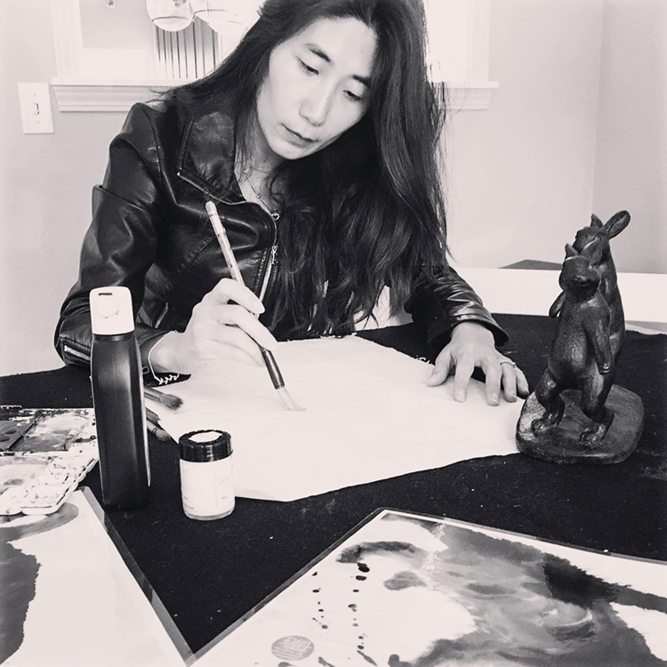 black-and-white portrait of artist Anita Wong, wearing a black leather jacket, drawing her paintbrush across paper