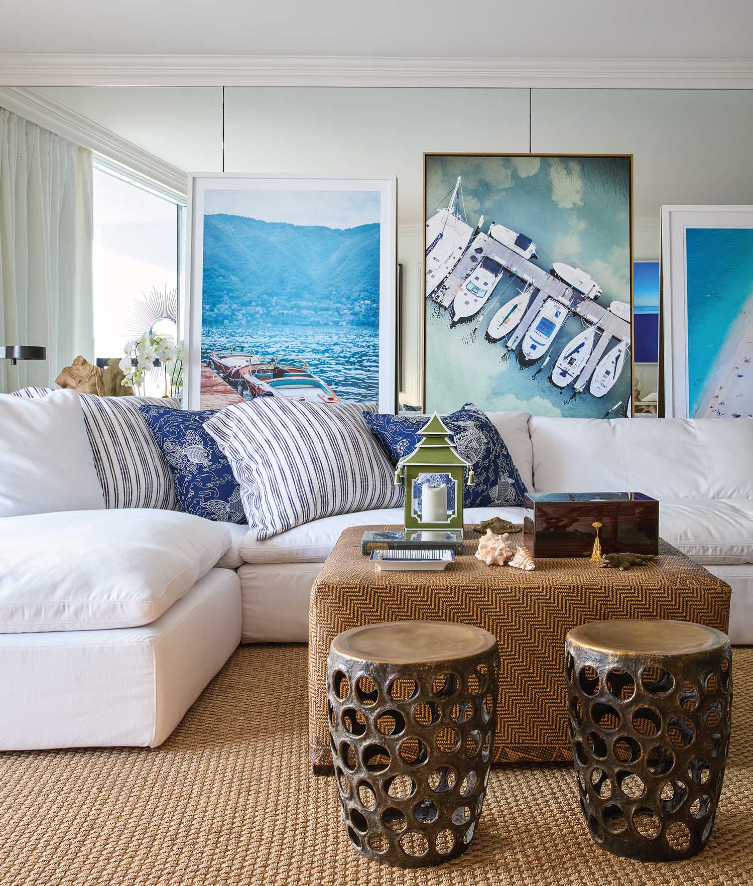 White sectional sofa with blue and white throw pillows and bronze stools. Boat and beach photos on the wall.