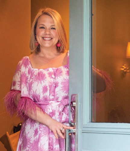 Rebecca Gardner greets party guests at the front door of a Savannah, Georgia, home. She wears a pink and white floral dress. The door is painted pale turquoise and features a large frosted window in the top half. 