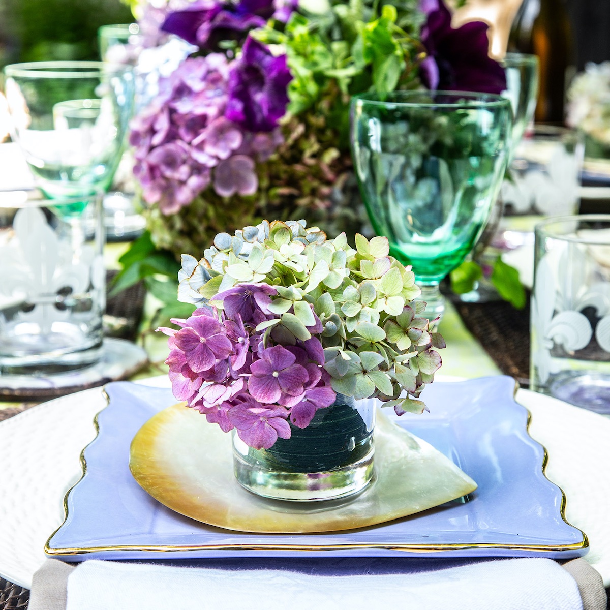 Single place setting with a disparate grouping of china and glassware, topped with a small arrangement of hydrangea blossoms in a glass tumbler.
