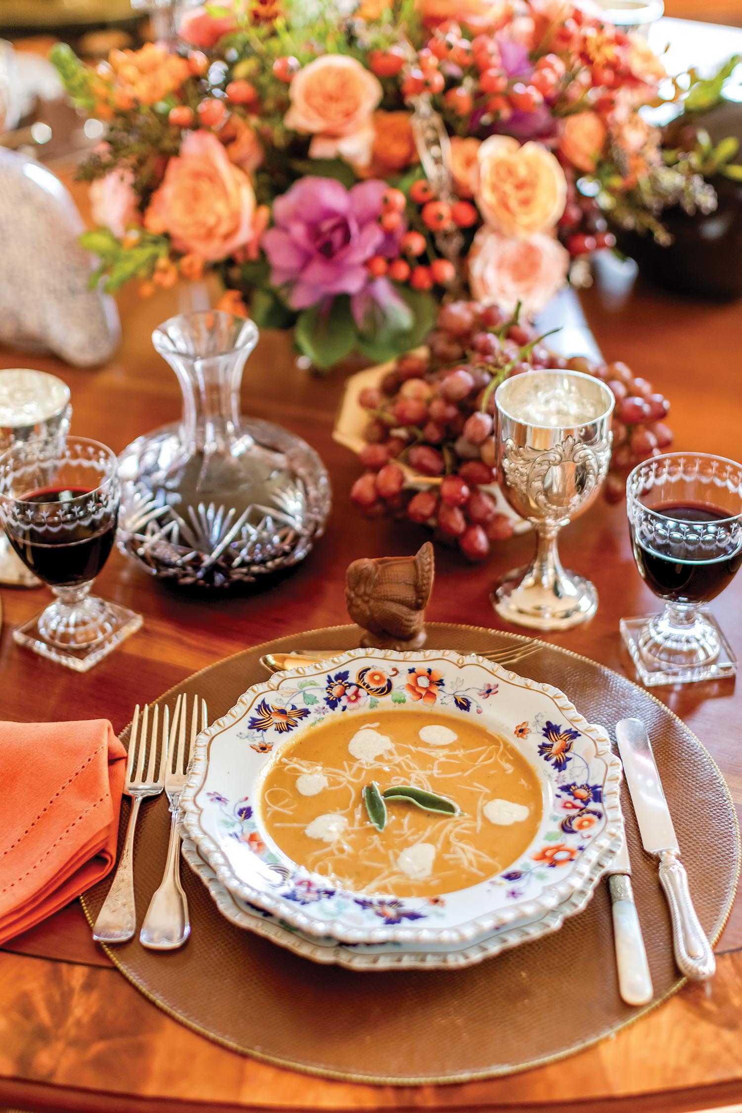 A bright gold soup sits in a painted floral bowl on a wooden table with orange and pink flowers.