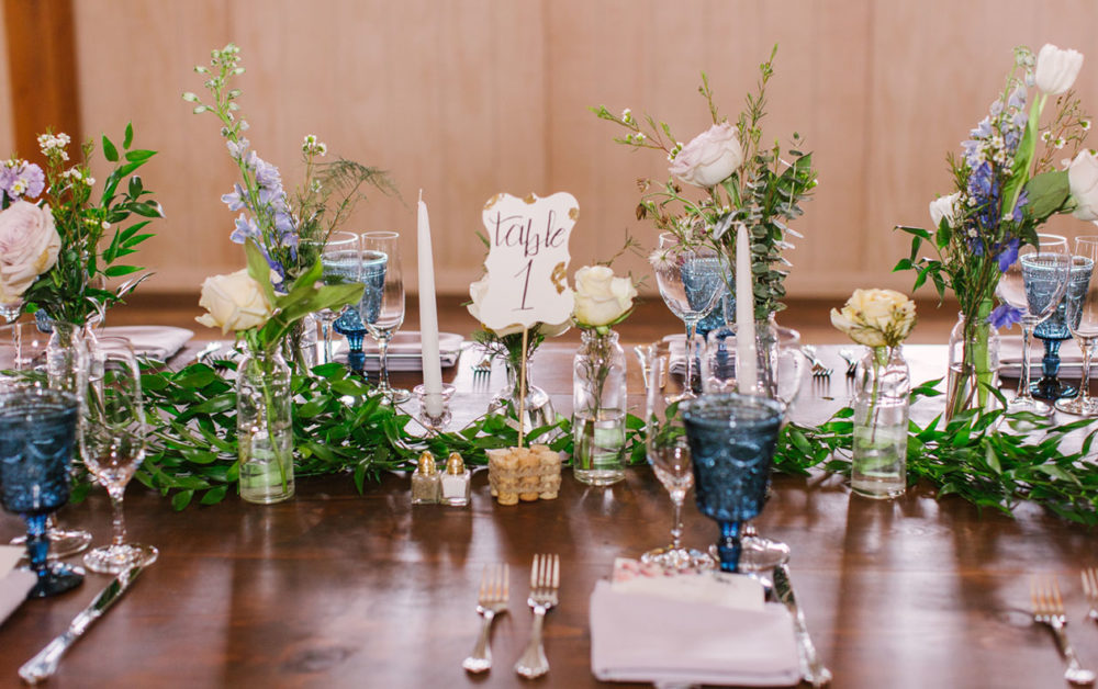 wedding table featuring flowers in small bottles scattered down the table, along with a runner of loose greenery