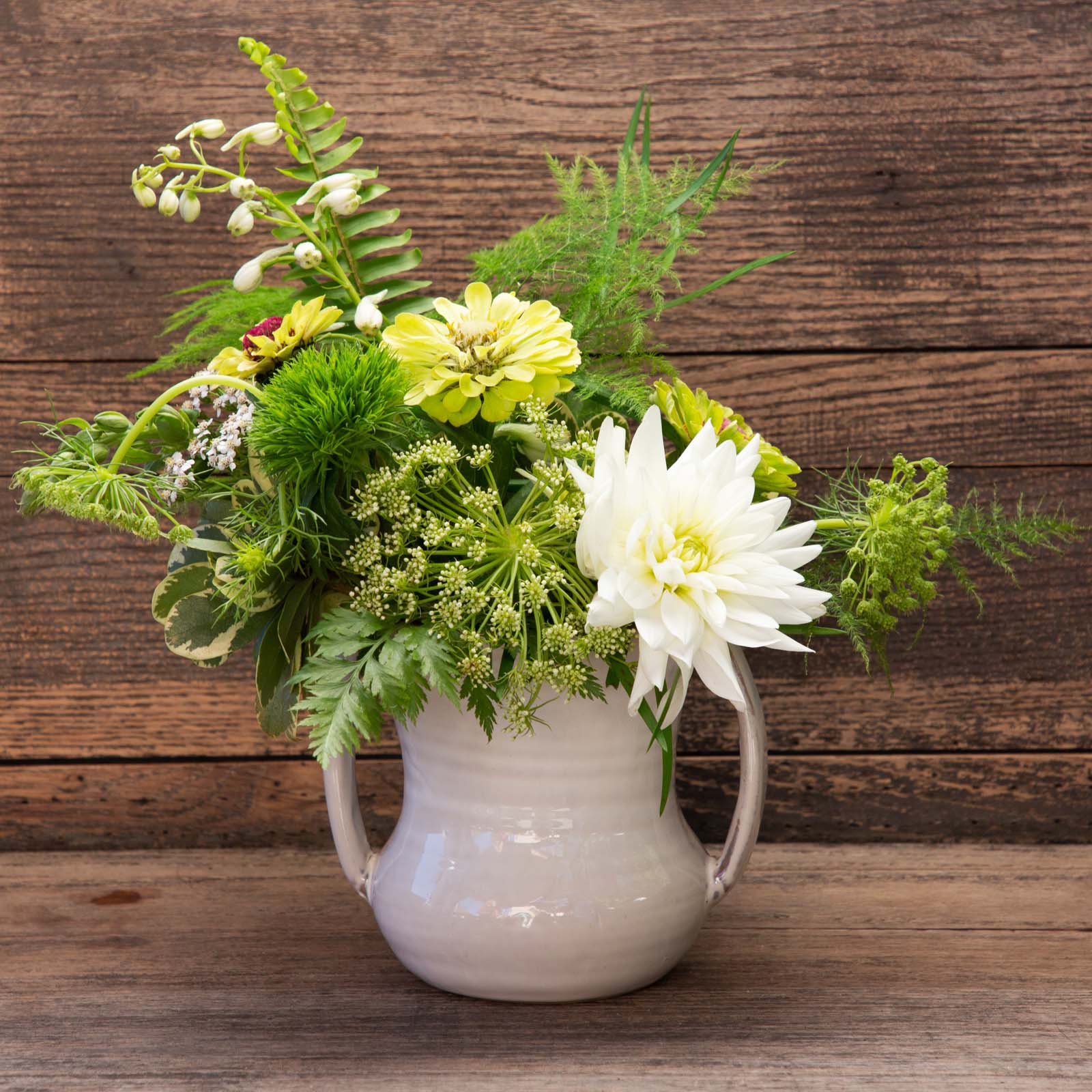White ceramic vase filled with green and white flowers (zinnias, dahlia, fern fronds and other foliage).