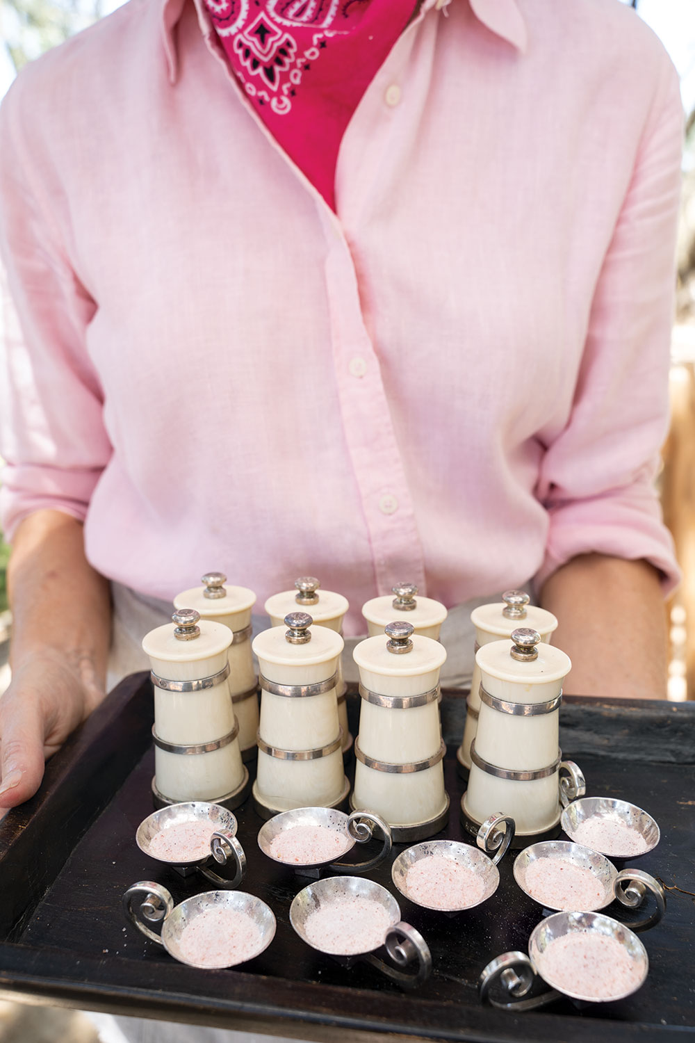 Woman in pink top with hot pink bandana holding a tray of silver salt cellars and pepper grinders