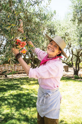 portrait of Frances Schultz clipping branches from her trees for a floral arrangement wearing a brimmed hat, neck scarf, and apron