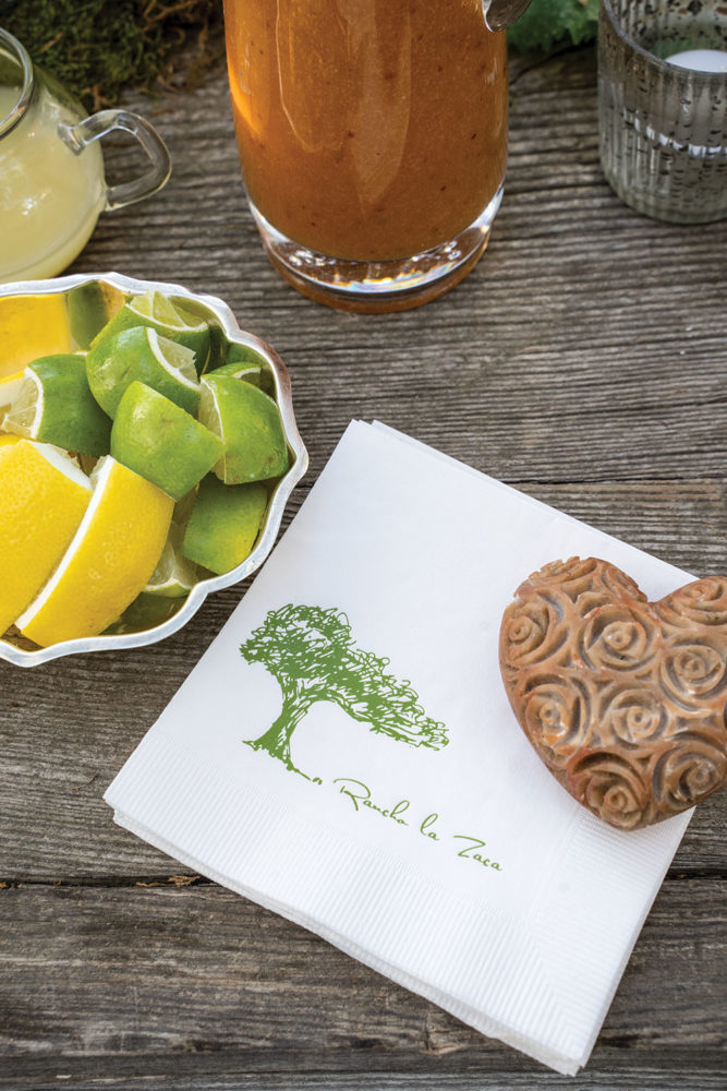 A stack of cocktail napkins bearing a tree motif and the name Rancho La Zaca beside a silver dish of sliced lemons and limes