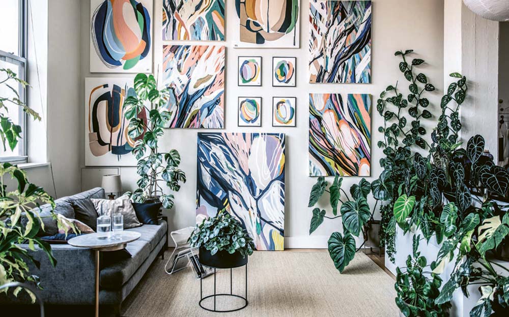A scene from the houseplant-filled apartment and studio of Brooklyn artist Alina Fassakhova. Her colorful, abstract botanical paintings fill the back wall. A charcoal-colored modern sofa sits under a window on the adjacent wall. The sofa faces a wall of lush, green plants; individual plants accent other areas of the room. 