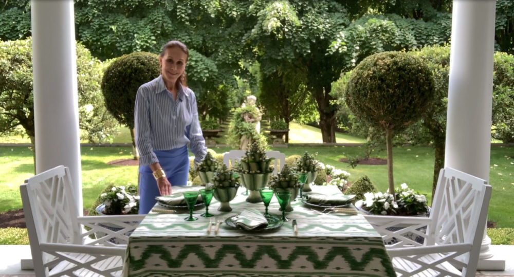 Carolyne Roehm stands beside an outdoor table set with green and white, with a view of a her formal garden behind her