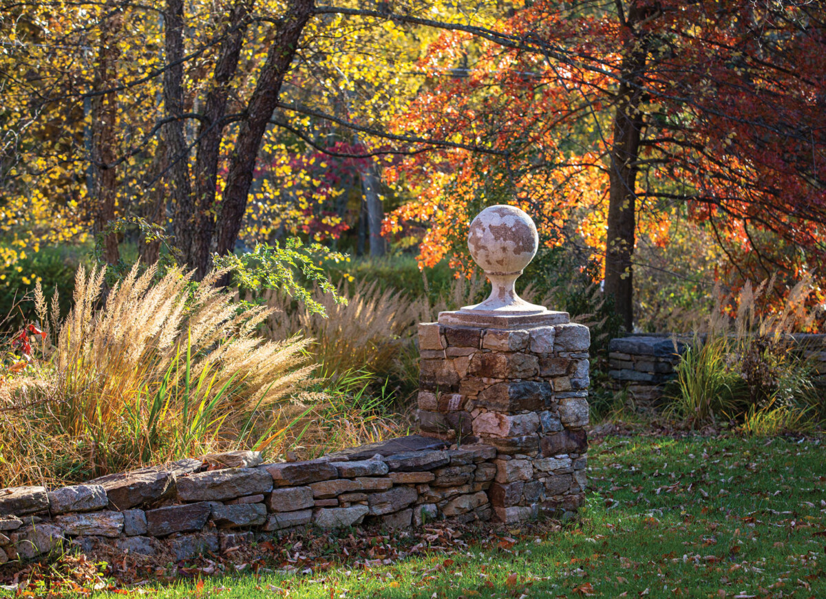 A low stone wall borders a bed of tall autumn grasses at Jardin de Buis in New Jersey. Sun shines through the red and yellow autumn foliage.