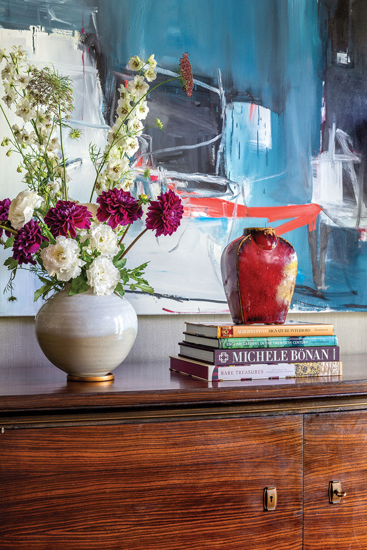 flowers, pottery vases, and a stack of books on a cleanl-lined wood sideboard. An abstract painting with blue tones and pop of red hangs in the background in a vignette by designer Matthew Patrick Smyth
