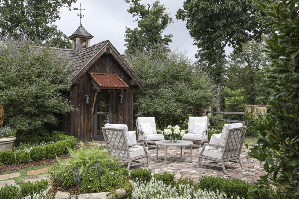 A set of 4 white-cushioned patio chairs, available through Blackjack Gardens in Birmingham AL, surround a small white table on a brick patio. The patio is surrounded by a lush green garden. The adjacent home is a rustic cabin with an aged metal overhang and a classic weathervane on roof.