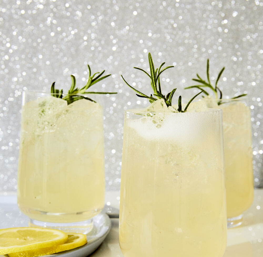 Three glass tumblers filled with Evergreen Sparkler, a fizzy lemon, rosemary, and gin cocktail from the book Merry Cocktails by Jessica Strand. Rosemary sprigs garnish each glass. The background is a glittery silver.