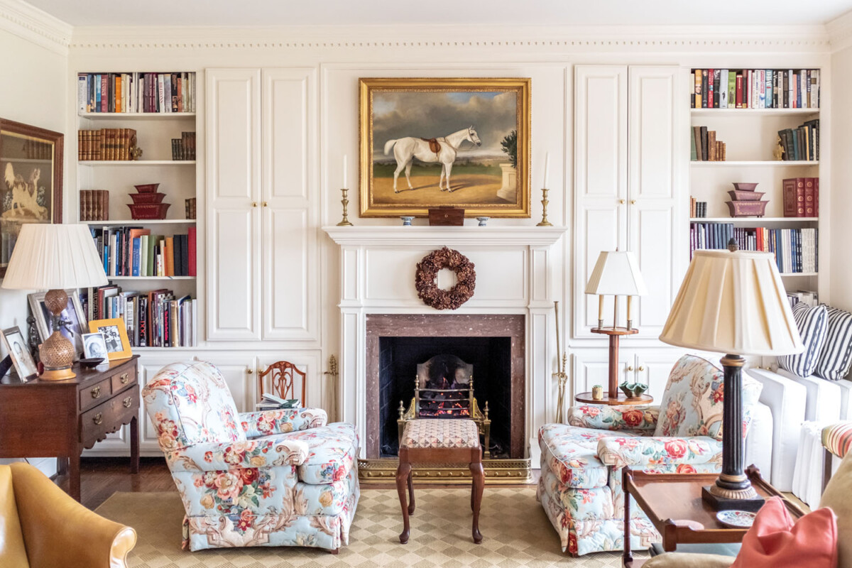 Living room scene with white paneled walls, a white mantel, flanked by built in book shelves and floral upholstered club chairs