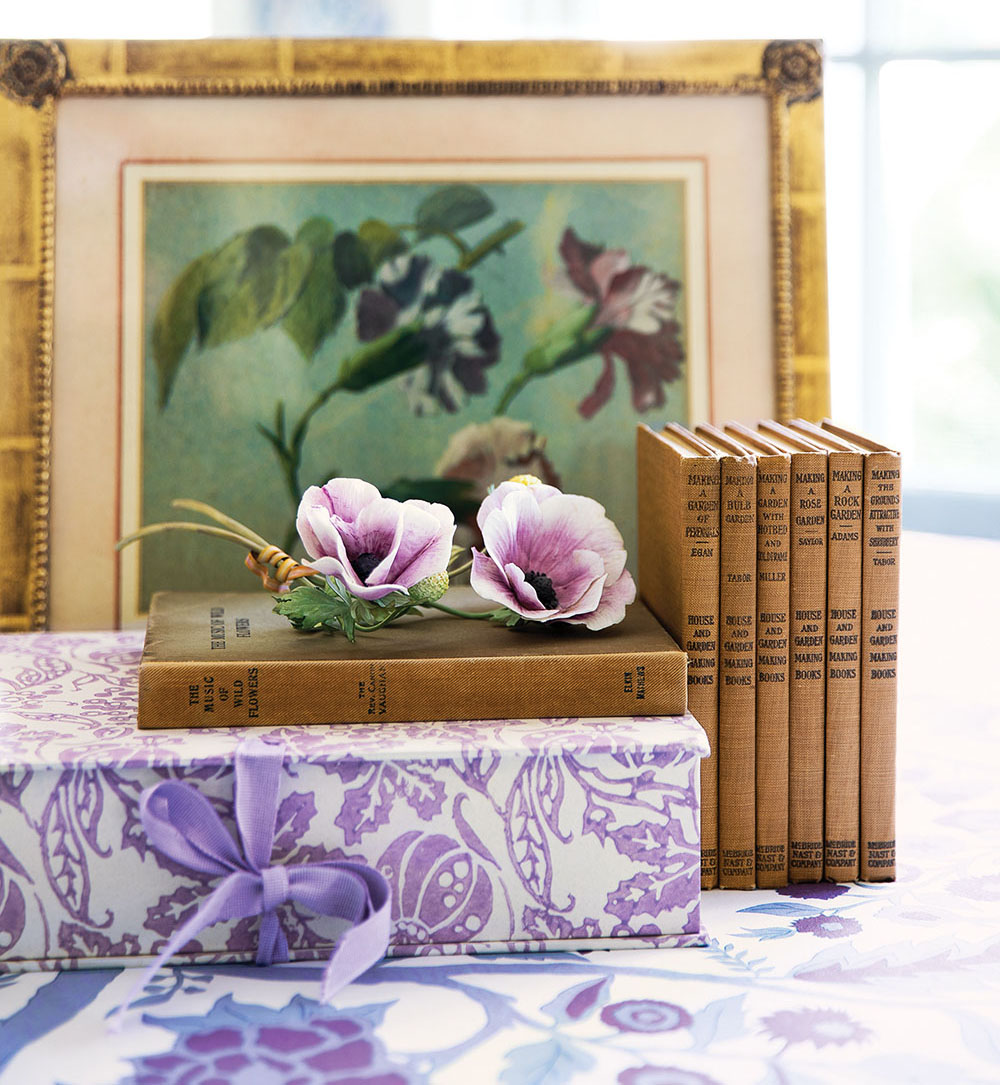 A vignette from the book Charlotte Moss Flowers including a framed floral painting, a set of vintage gardening books, two purple blooms, a present wrapped in a purple-on-white floral paper, all arranged on a purple-on-white floral print fabric