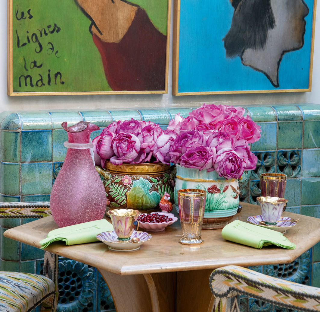 A table setting beside a blue and green tiled wall at the Parisian home of Terry de Gunzburg. Garden roses tinged bright pink fill a pair of colorful porcelain vessels bearing floral motifs. The setting also features artful glassware (pitcher, drinking glasses), china teacups, and a serving dish of pomegranate seeds, all carrying through the pink color scheme. Pale green napkins contrast with the pink and tie into the blue and green featured in the tile wall, artwork, and chair upholstery