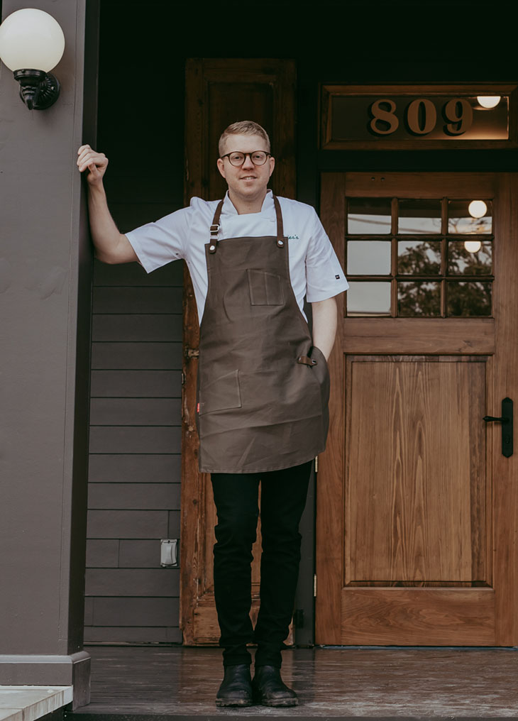 portrait of of chef Hunter Jackson, wearing an apron, white shirt, and black pants, standing on the front porch of his restaurant Elvie's in Jackson, MS