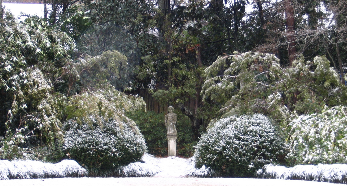A circular lawn covered in snow winter know, surrounded by garden beds and taller shrubs beyond. A statue of a child, standing on a pedestal where the pushes part, is the focal point. Garden design by Mary Walton Upchurch