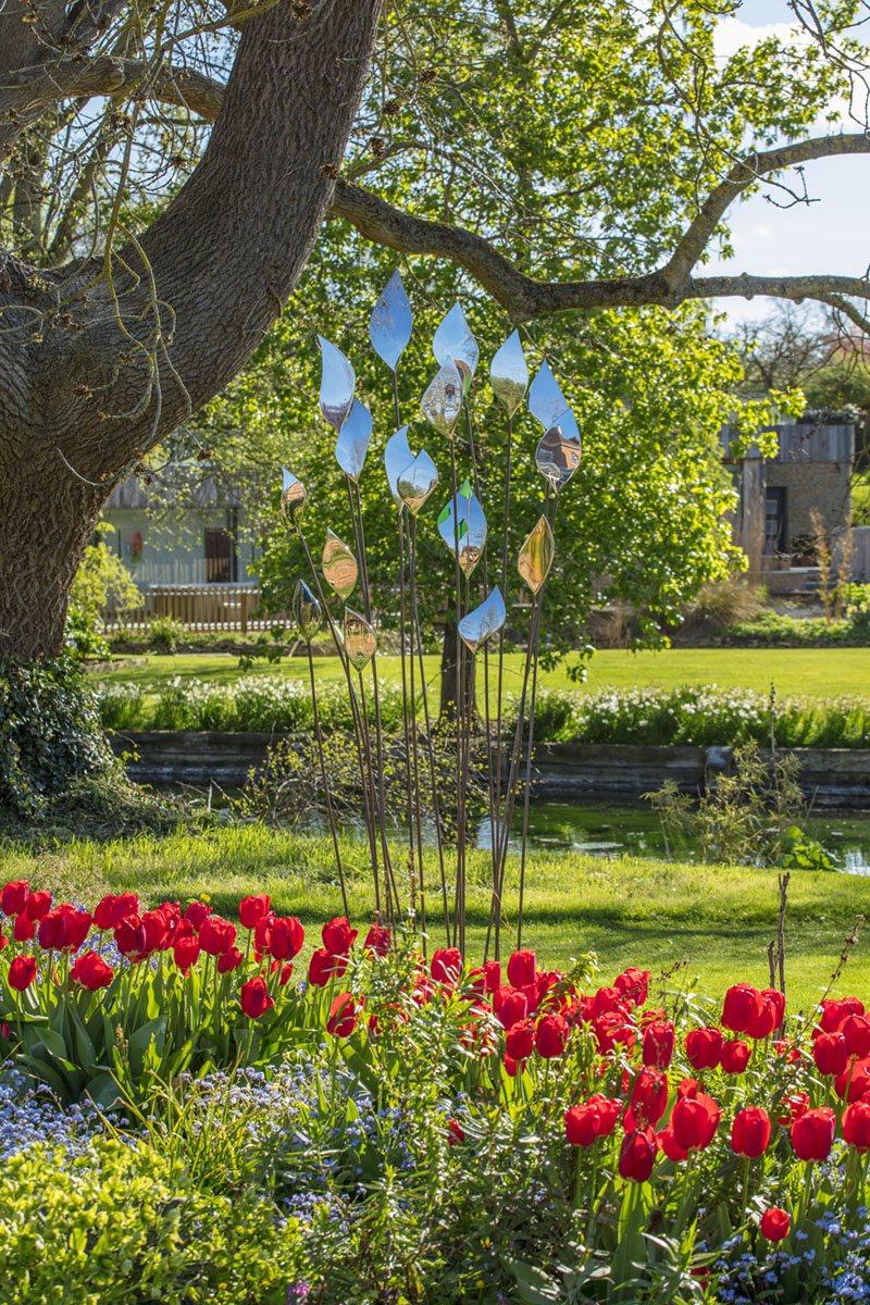 David Harber Sculpture metal leaf sculpture in a bed of red tulips in a garden with a pond and large oak tree