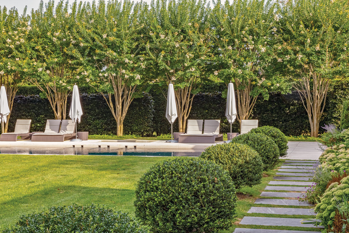 A straight path of rectangular stepping stones, bordered by rounded boxwoods, leads to a pool lined by a row of crepe myrtles, landscape architecture by Quincy Hammond