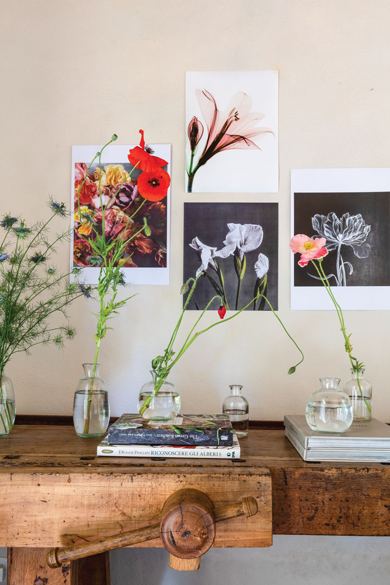 A vignette at PUSCINA FLOWERS includes simple unframed prints of flower photography on the wall, small bottles filled with poppies, and books on an antique wooden table