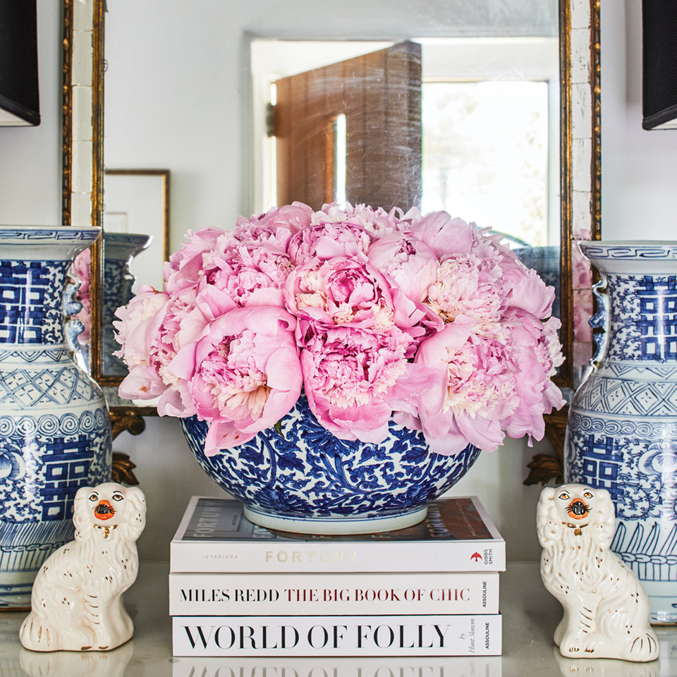 A decorative vignette in Gavin Duke's foyer features pink peonies filling a blue-and-white porcelain bowl, sitting on a stack of books. A pair of porcelain dogs and blue-and-white vases flank either side. In the background is an ornate gilt mirror. The reflection reveals a crystal chandelier and an open, natural wood front door. the walls are painted a soothing cool white.