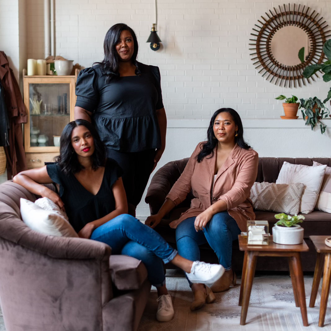 The team of The Wild Mother floral and event design studio. Callie and Leah Palmer sit on a pair a cattycorner sofas in their studio. Lauren, wearing black, stands between them.
