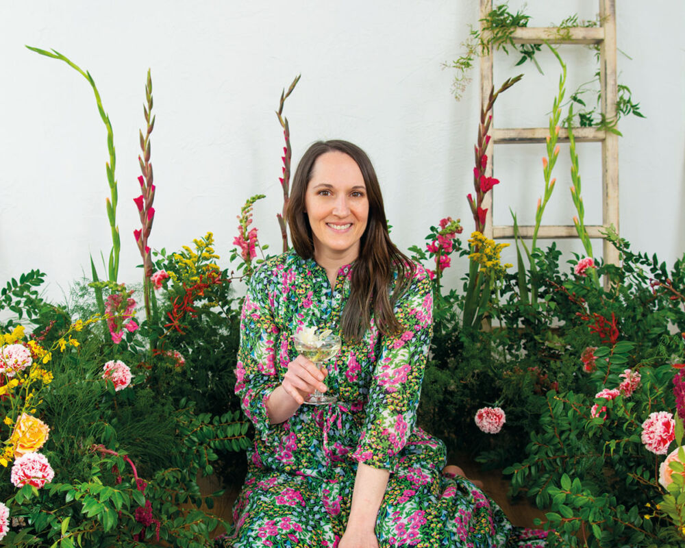 portrait of Alyson Brown, author of The Floral-Infused Cocktail, wearing a floral dress, surrounded by a lush floral installation, holding a cocktail in her hand