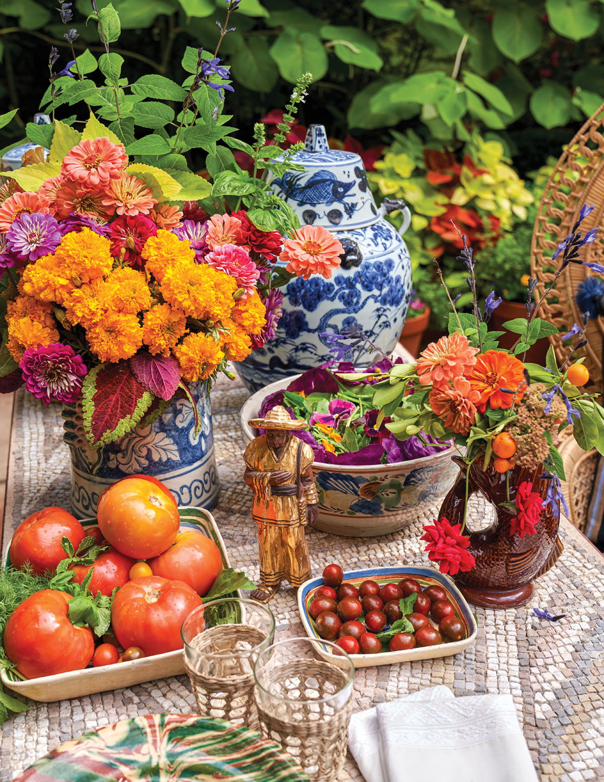 A blue-and-white ginger jar, a large salad, shallow bowls of summer tomatoes, and a pair of lush red and orange floral arrangement fill a mosaic patio table