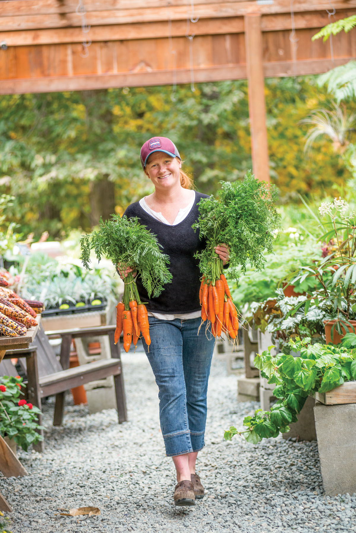 Allie Boeri, wearing her red hair in a ponytail under a ballcap, walks through the open-air farmers market carrying a bunch of carrots in each hand