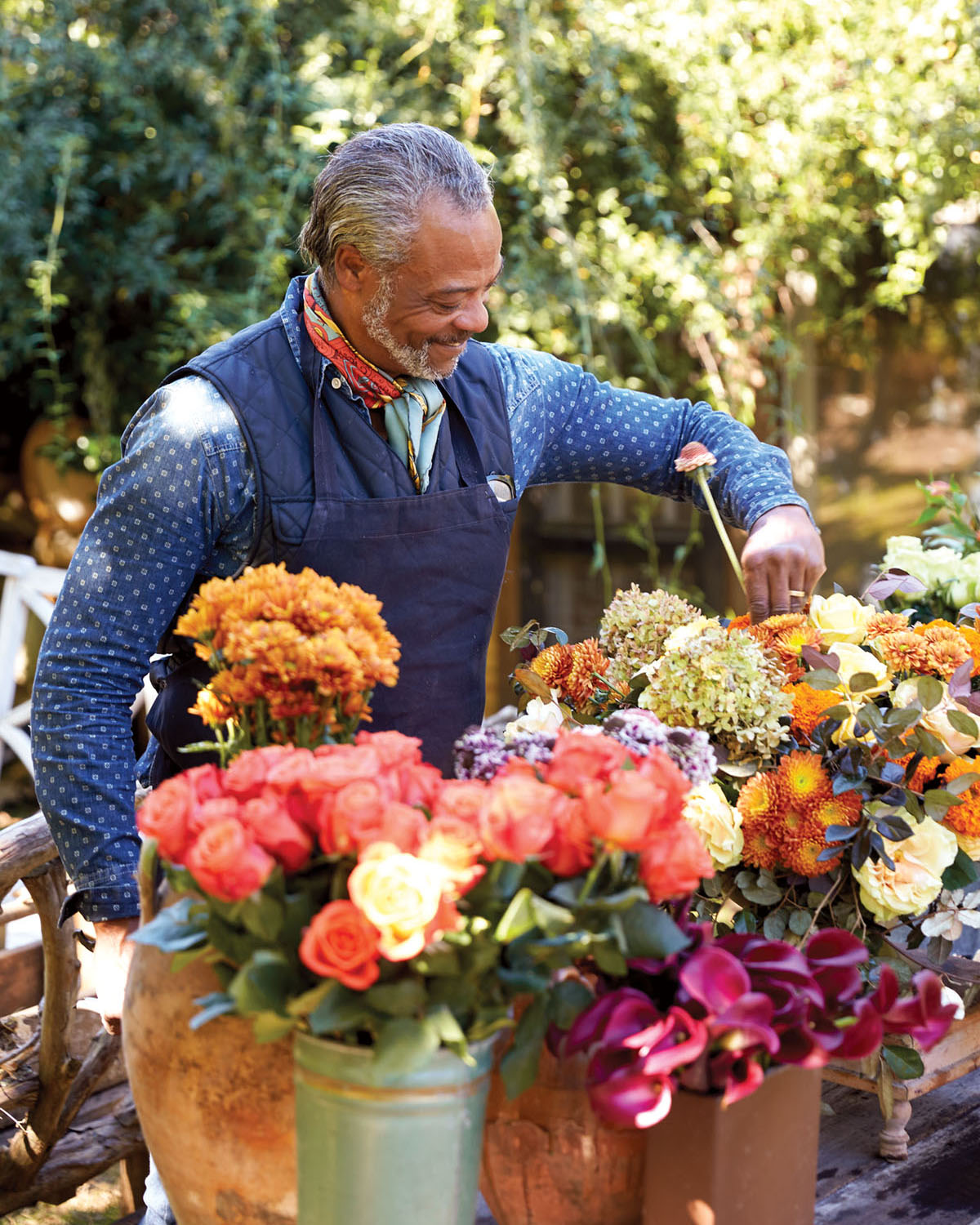 Keith Robinson, wearing a long-sleeved denim shirt, vest, and bandana around his neck, arranges fall flowers in a variety of coral and orange hues