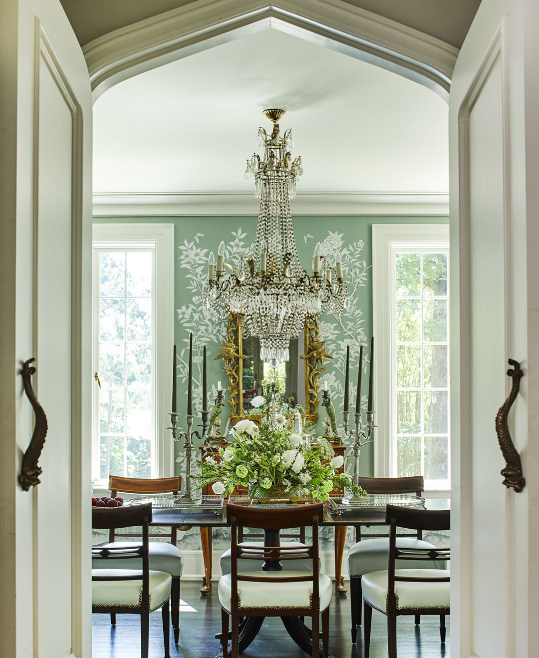 Double doors open to reveal a dining room papered in a custom, soft blue-green Gracie wallpaper, with an crystal chandelier and ornate gilt mirror