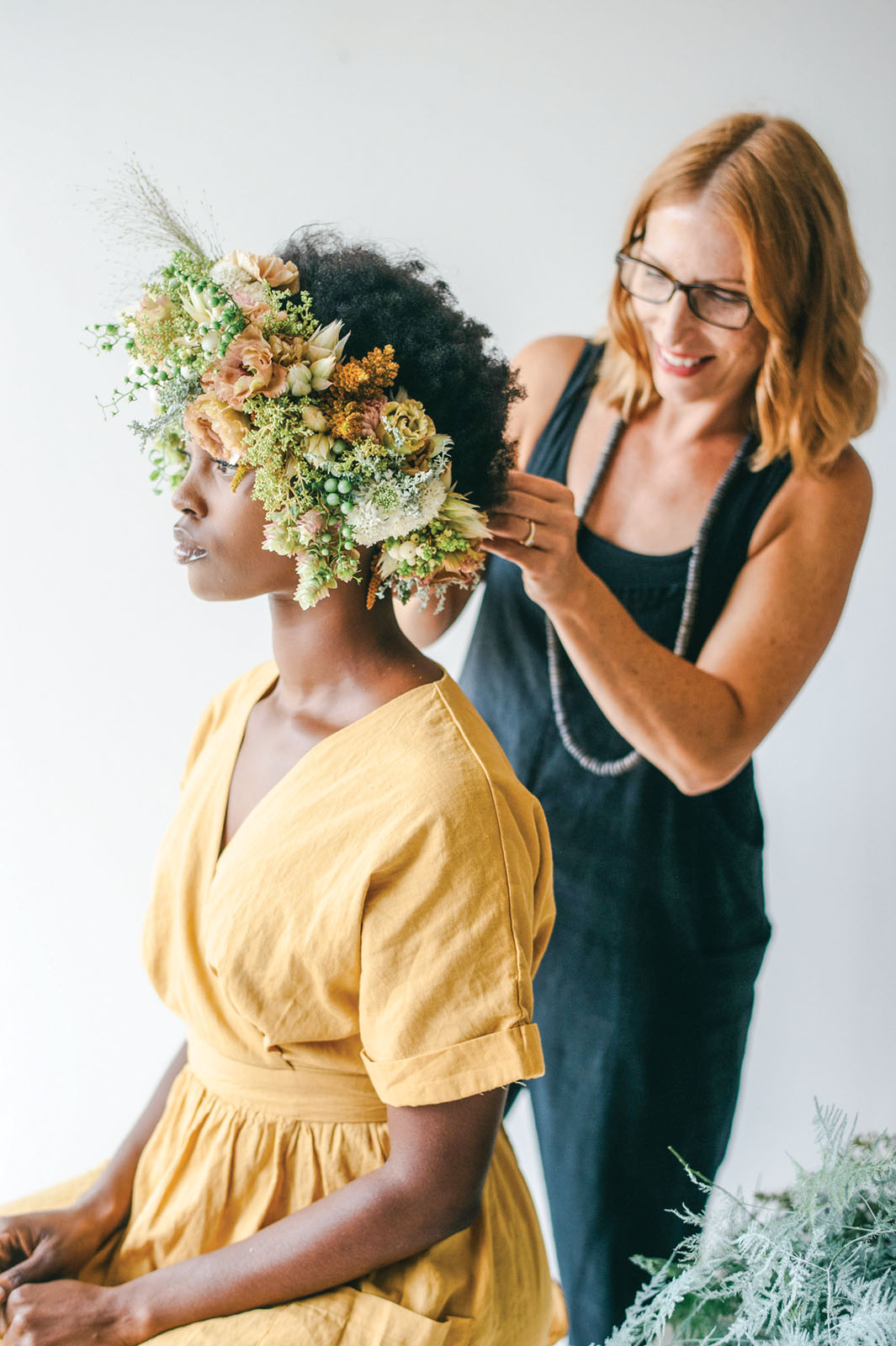 portrait of floral designer McLeary, wearing a black tank top, styling a floral headpiece on a model, who wears a yellow dress