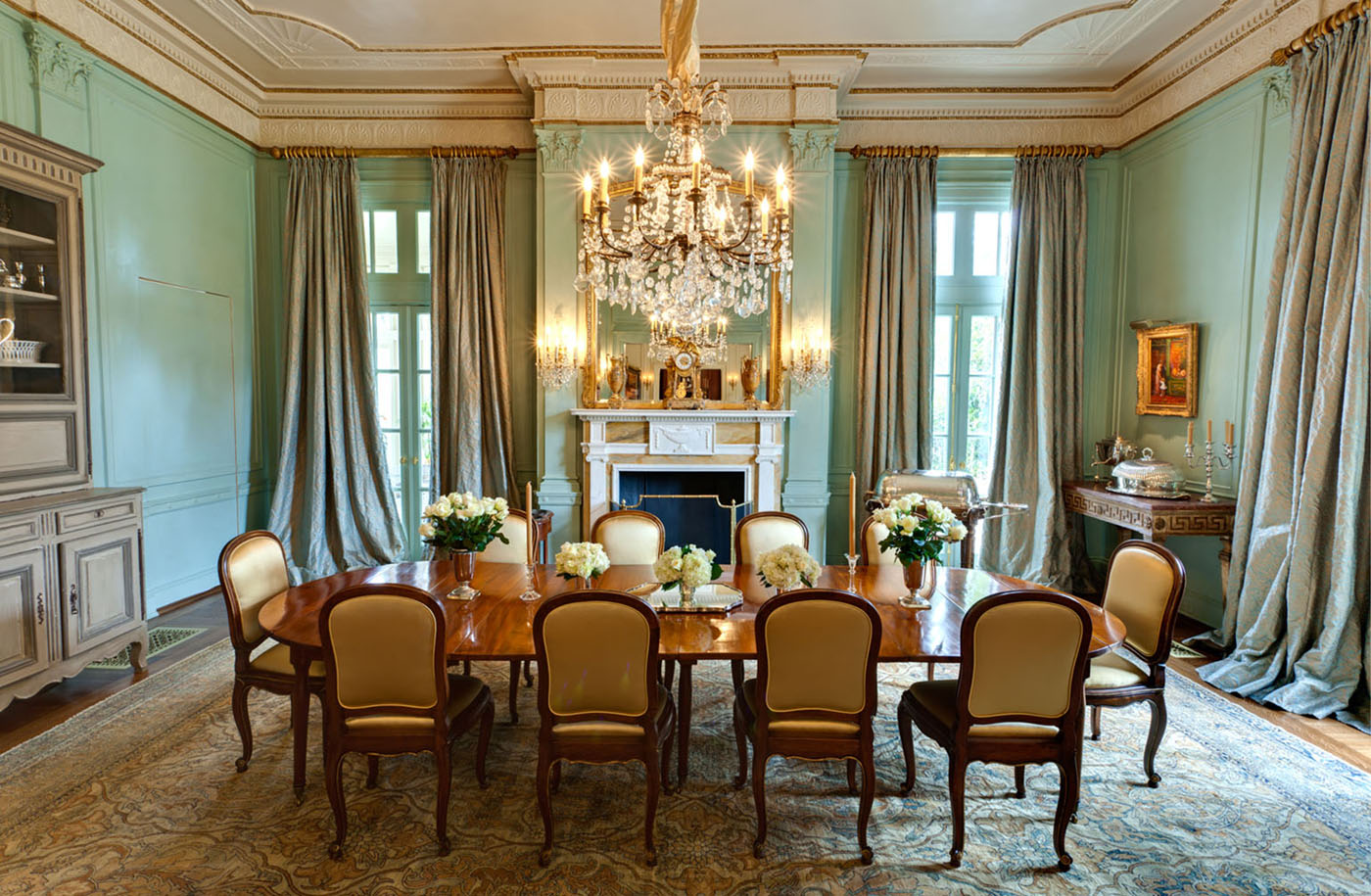 An elegant Robin's egg blue dining room with floor to ceiling taupe drapes, a crystal chandelier an antique rug in muted colors featuring an oriental rug pattern