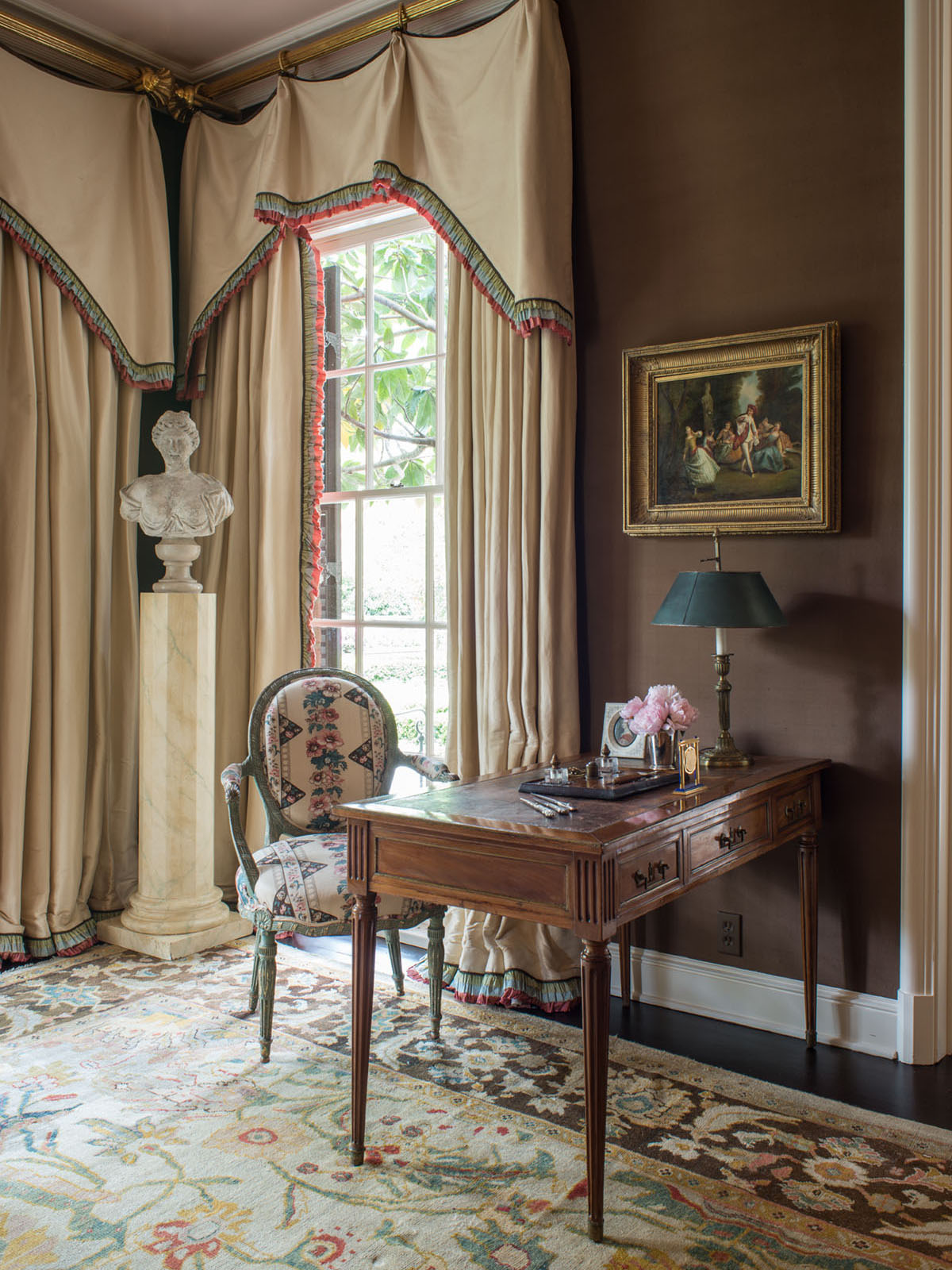 A study painted deep taupe features an antique desk and chair, a carved bust displayed on a pedestal, tailored valances and floor-to-ceiling drapes, and a Persian Sultanabad floral rug pattern, featuring yellow, coral, and green motifs on an cream background and dark brown border.