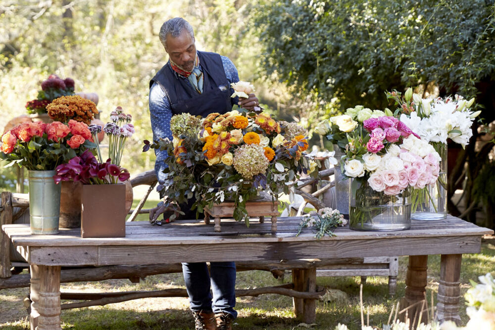 Keith Robinson standing behind antique table in garden, creating flower arrangement.
