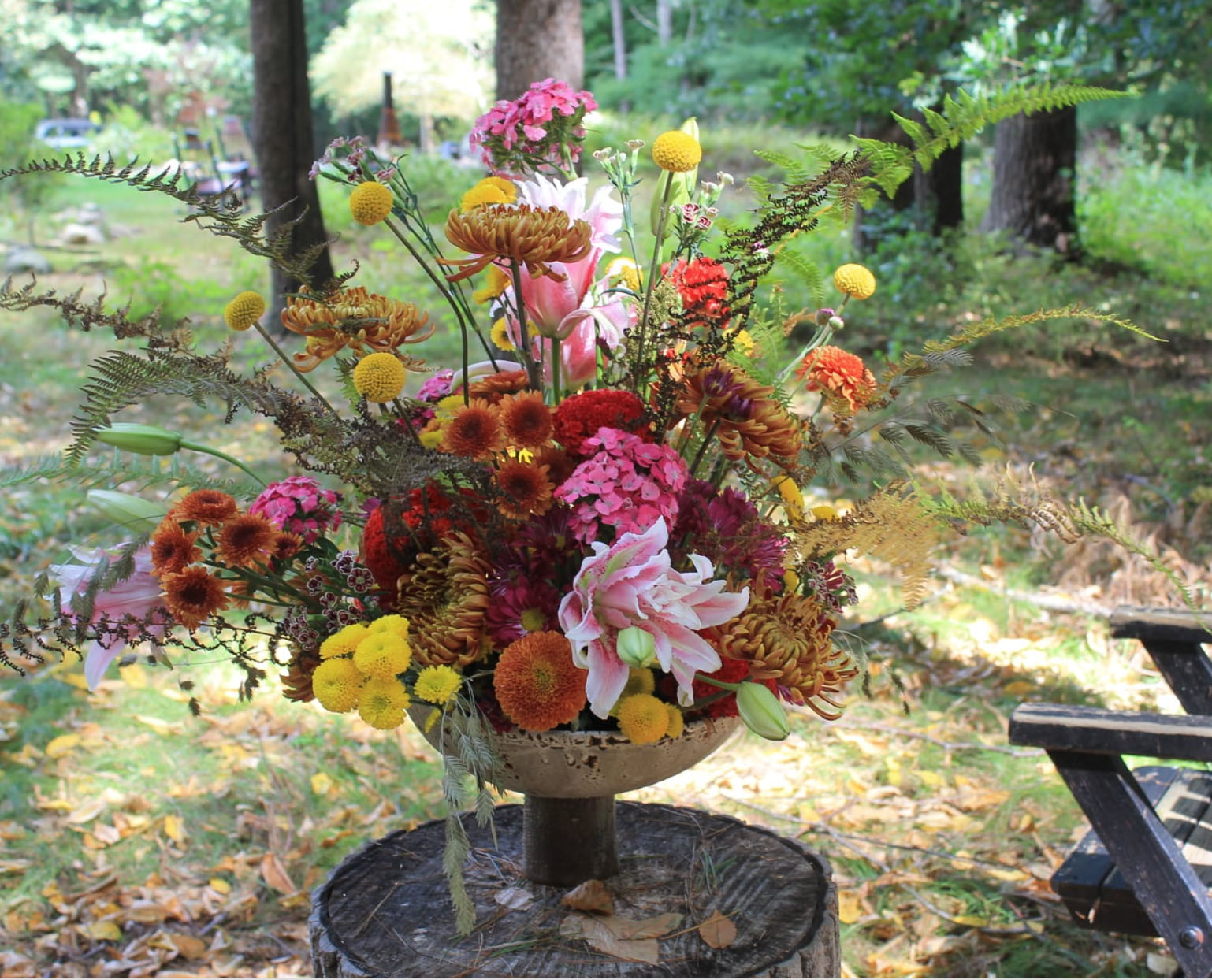 Fall orange and pink flower arrangement in footed compote atop stump in garden.