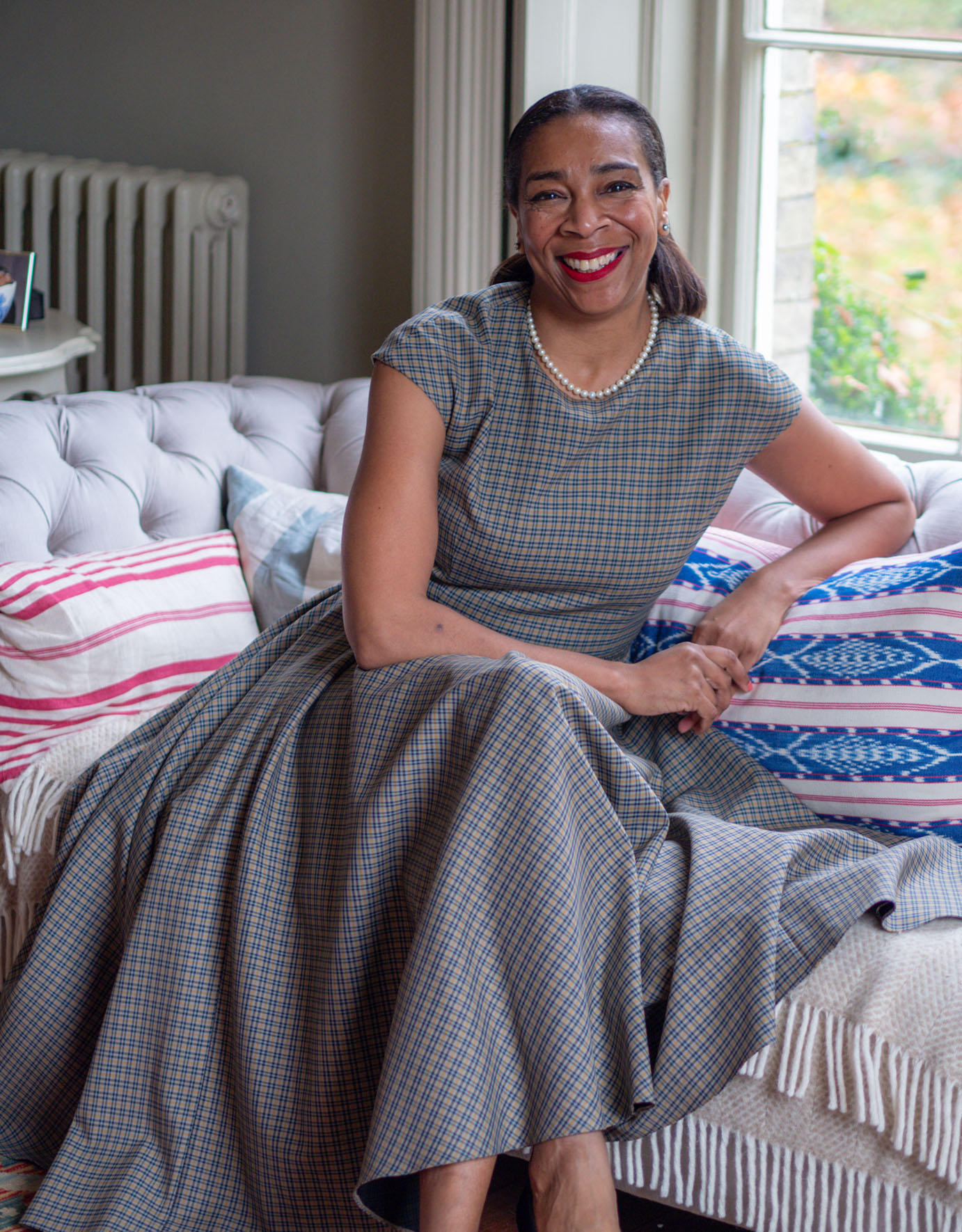 Paula Sutton, creator of Hill House Vintage, poses on a sofa at home, wearing pearls and a 1950s retro-style full-skirted gray dress and pearls