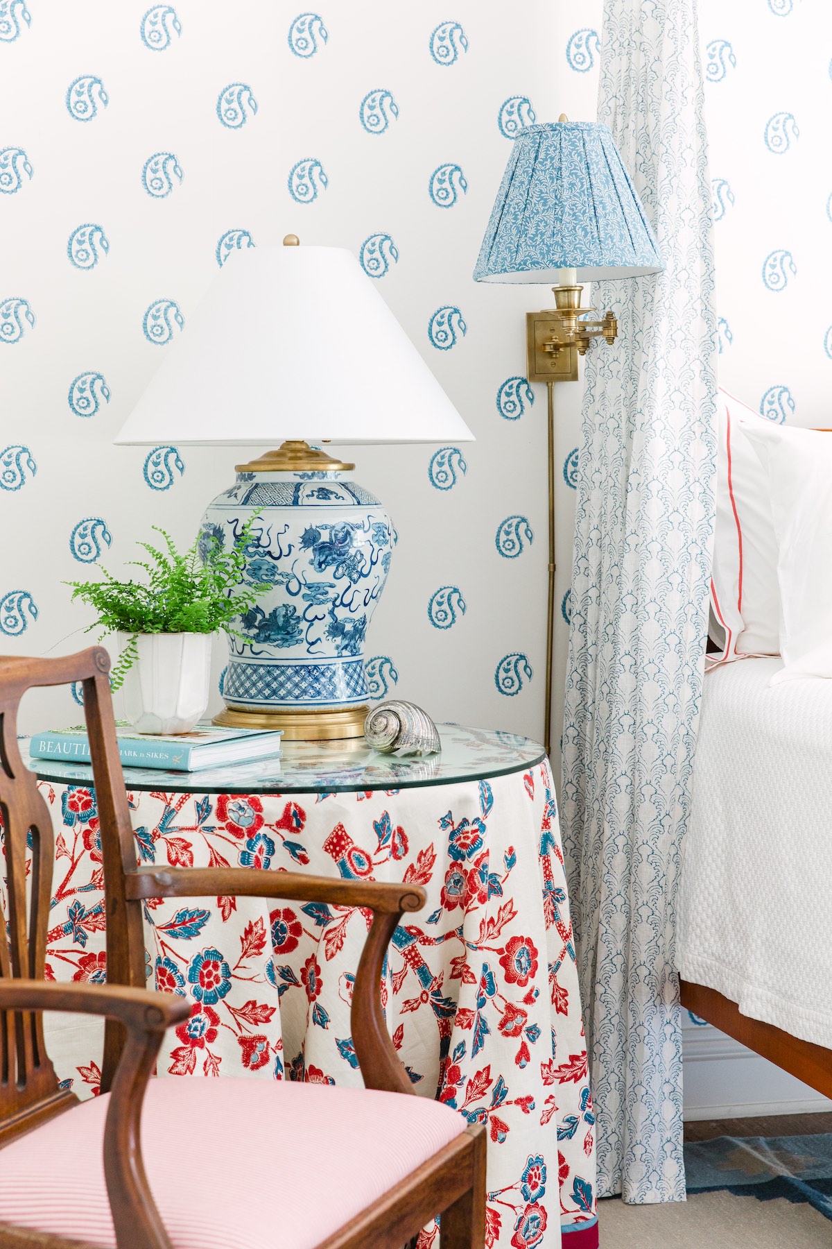 Bedside table with floral print table cloth, blue and white jar lamp and potted fern. Brass swing-arm lamp with pleated blue and white shade mounted on wall beside bed.
