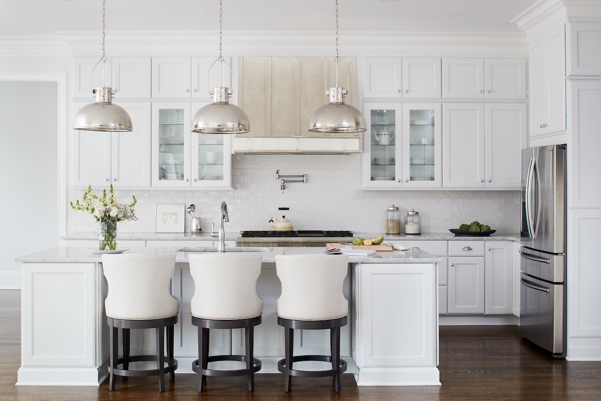 White kitchen with three stools at island, bright hammered nickel pendants over island, and light rail moldings hiding under-cabinet LEDs above countertops.