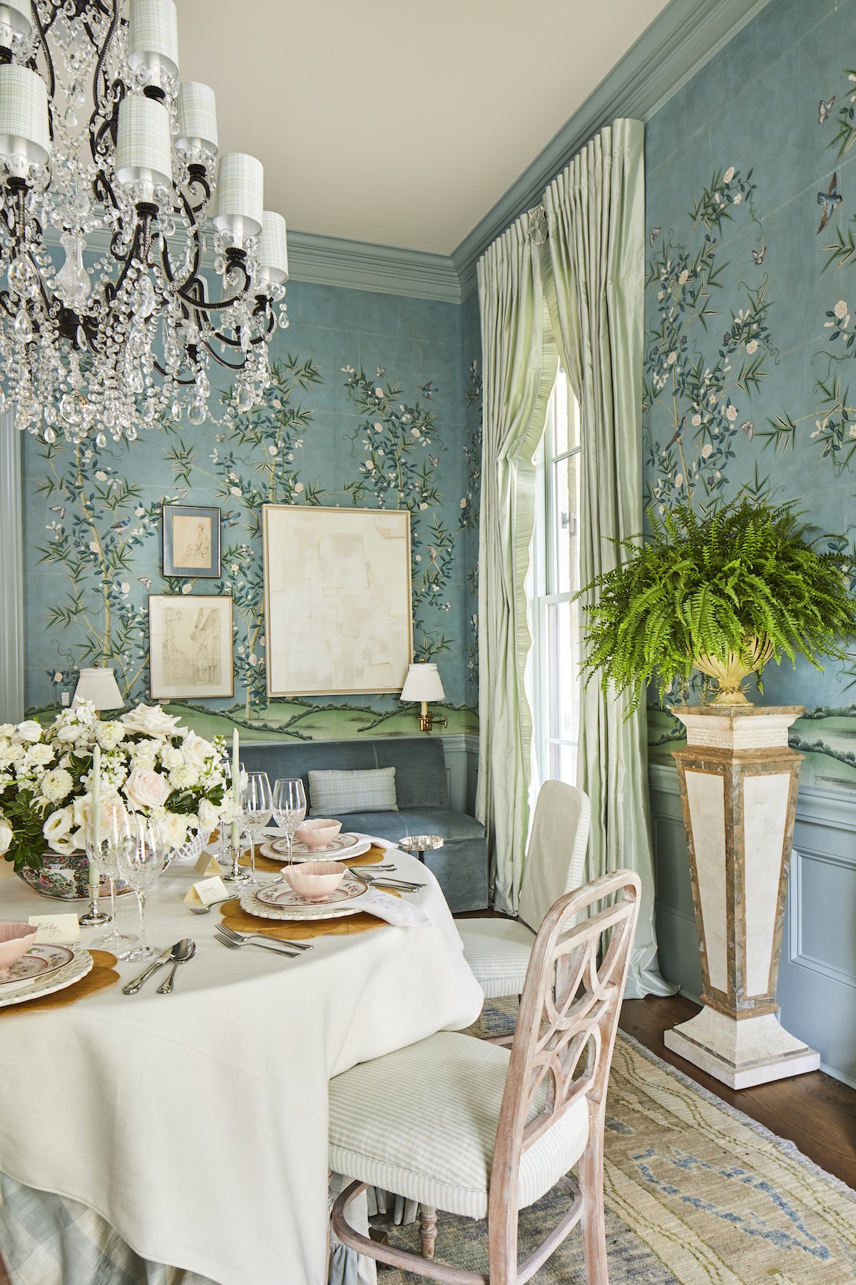 Dining room with table set with china and white flower arrangement. Blue banquette with a pair of brass sconces sits against the wall behind the table.